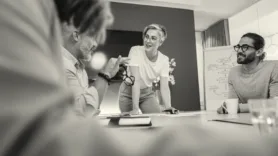 Several coworkers having a discussion around a table, with a whiteboard in the background