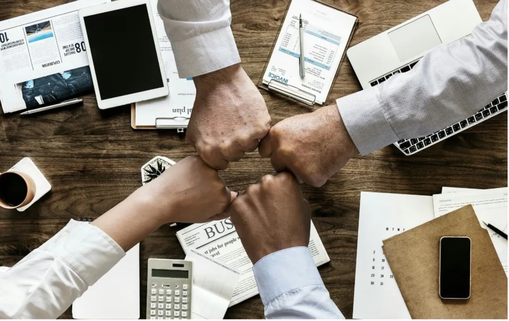 four hands in fists, forming a square above a desk strewn with papers and devices