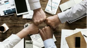 four arms meeting in a fist bump over a table with papers, laptops, and calculcators
